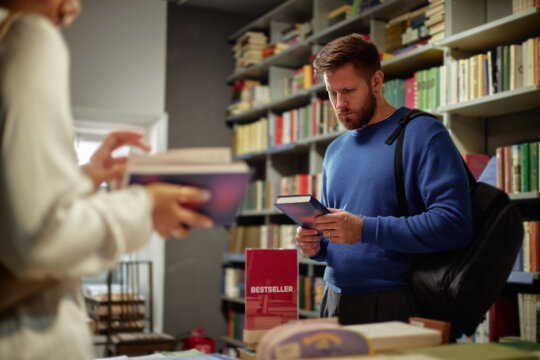 Caucasian young adult man browsing books in bookstore, holding book and reading cover while standing near shelves, another with book visible in foreground, books filling background