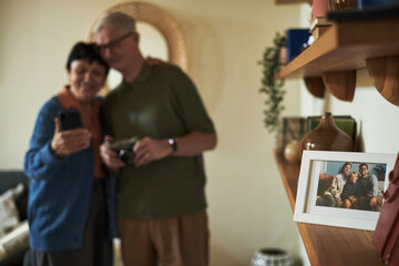 Senior couple standing closely, smiling, and posing with smartphone in cozy living room. Background includes framed photo on shelf and warm home decor creating a welcoming environment
