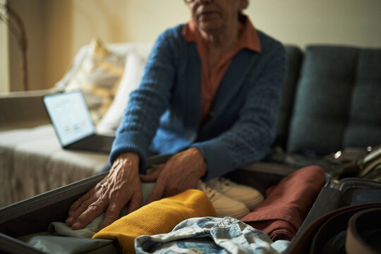Elderly woman packing clothes into suitcase with open laptop on bed showing serene and organized environment for traveling moment
