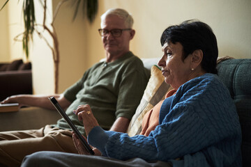 Elderly couple relaxing on couch holding digital tablet in warm-lit living room. Woman in blue cardigan engaging in conversation with man wearing glasses