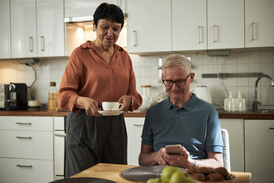 Elderly couple sitting in modern kitchen, man using smartphone while woman smiling and holding cup of tea. Atmosphere of warmth and tranquility surrounding them