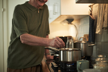 Elderly man smiling and stirring a pot on stove in cozy home kitchen. Warm lighting and utensils create inviting atmosphere, highlighting enjoyment of cooking