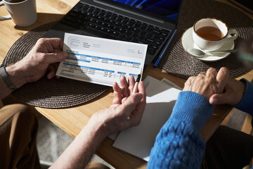 Elderly couple sitting at a table holding hands and reviewing financial documents and utility bills, with laptop and coffee cup visible in the background