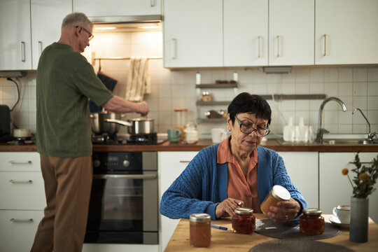 Senior couple making homemade preserves in modern kitchen Senior woman looking at jars with jam while senior man is stirring pot on stove