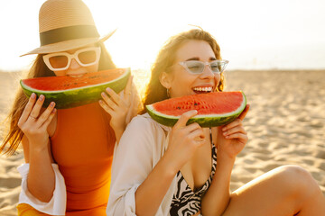 Happy female friends having fun and eating watermelon on a sunset beach. Two beautiful women...