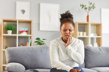 Young African American woman with caries sitting on sofa on living room background at home, holding...