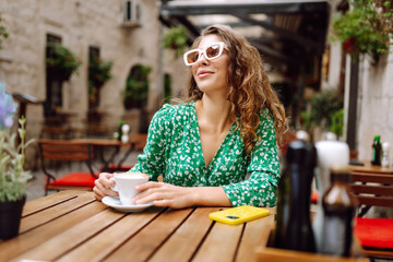 Portrait of happy woman drinking cup of coffee at wooden table on cozy veranda. Young tourist enjoying morning and hot drink outdoors. Tourism and drinks concept.