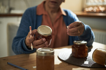 Elderly woman holding jar of homemade preserves while sitting at table in cozy kitchen. Light streaming through window highlighting the jars