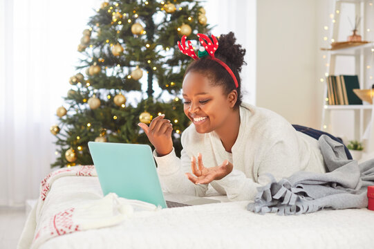 Cheerful young African American woman lying on bed with laptop, making online video call during Christmas Eve. Positive black girl celebrating x-mas at home, congratulating friends or relatives online - Powered by Adobe