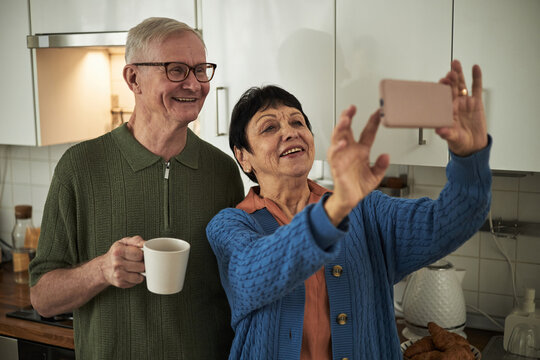 Smiling elderly couple taking selfie in modern kitchen while holding coffee mug, capturing cheerful moment and engagement in selfie activity, adding warmth to home setting