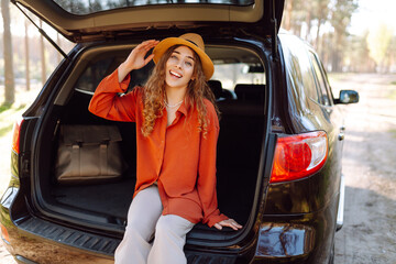 Young woman sits in the trunk of a car and enjoys the weather in a sunny forest. Traveler on a car...