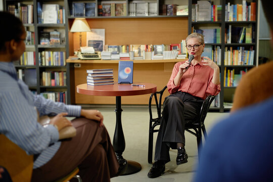 Senior Caucasian woman sitting on chair speaking into microphone gesturing with hand during session in bookstore with audience listening and books displayed on table - Powered by Adobe