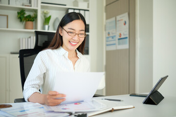 Smiling businesswoman wearing glasses reading documents while enjoying a cup of coffee in modern office.