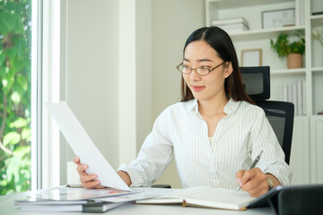 Focused businesswoman wearing glasses reviewing a document and writing notes while working at her office desk.