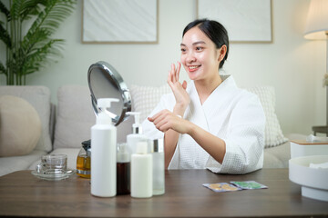 Happy Asian woman applying facial skincare in a bright living room. Wellness and Self care concept