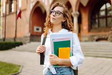 Beautiful student with glasses and books in hands stands against university campus background. Nice woman with stack of books and notebooks enjoying weather at sunset. Education concept. Lifestyle.