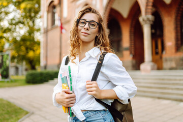 Beautiful student with glasses and books in hands stands against university campus background. Nice woman with stack of books and notebooks enjoying weather at sunset. Education concept. Lifestyle.