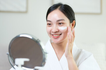 Smiling Asian woman in a bathrobe enjoying her beauty ritual, gently spreading face cream in a ...