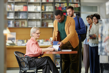 Caucasian woman sitting at table signing book for Black man while diverse group of people standing...