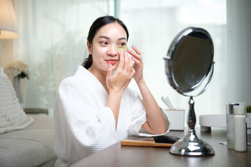 Young Asian female applying eye patch for hydration in a cozy room. Self care concept.