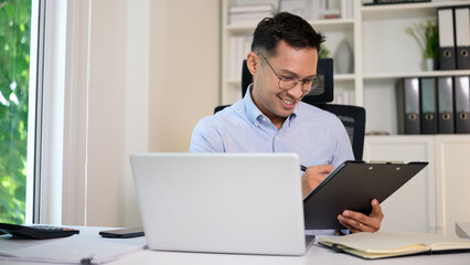 Smiling male professional writing notes on a clipboard while working at his desk. Bright and positive office setup with laptop and natural light.