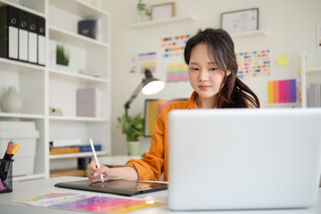 Young female graphic designer using a digital pen tablet and laptop to create artwork in a bright workspace.