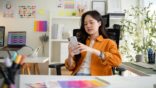 Young female graphic designer browsing smartphone in a stylish, plant-filled workspace. Taking a break from creative tasks.