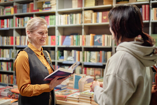 Middle aged Caucasian woman smiling while holding open book and talking to young adult woman in bookstore, bookshelves filled with colorful books visible in background