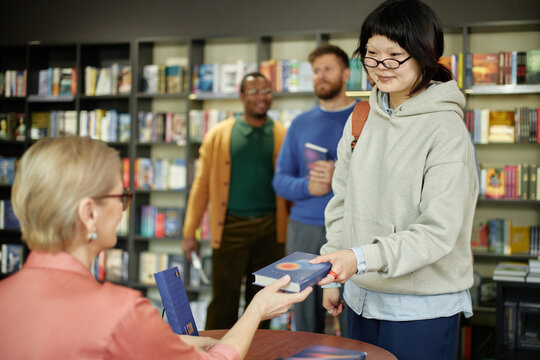 Asian young adult woman receiving book with autograph from Caucasian female author during session, multiethnic group of young adult men standing in background holding books - Powered by Adobe