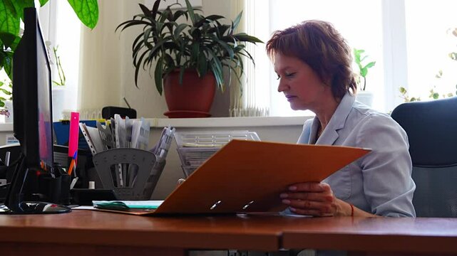 Woman working with folders and documents at her desk in an office setting. Paperwork routine, archive management, and administrative desk tasks.