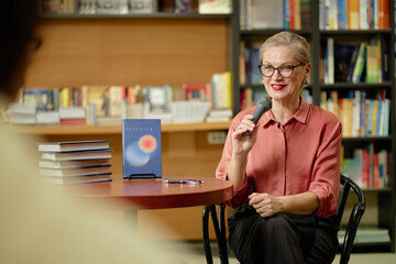 Caucasian senior woman holding microphone speaking at book event in bookstore, stack of books and...