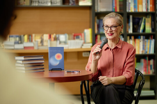 Caucasian senior woman holding microphone speaking at book event in bookstore, stack of books and pen on table, bookshelves in background, eyeglasses and short hair visible