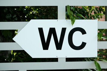 White directional sign with black WC letters pointing left, mounted on a fence with green leaves in...