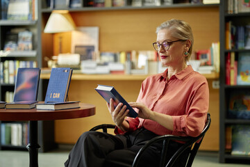 Caucasian senior woman with short gray hair and glasses sitting in chair reading book in library,...