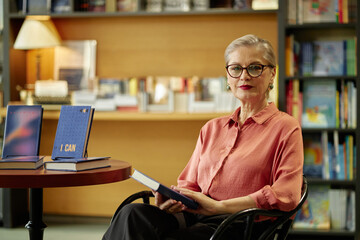 Portrait of senior Caucasian woman with short gray hair and glasses sitting in library holding...