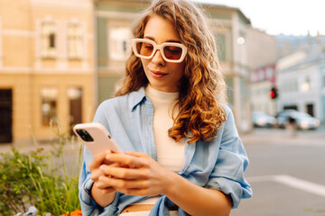 Happy woman in sunglasses and with phone walking along sunny city streets. Young woman blogging and...