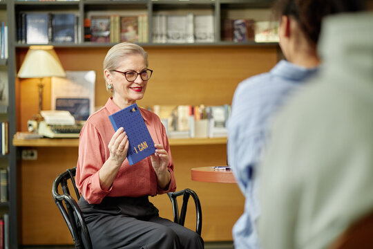 Caucasian senior woman wearing glasses holding motivational book while sitting in library engaging with young adult man during session