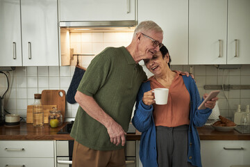 Elderly couple standing in kitchen, smiling while having morning coffee and using smartphone. Senior lady holding mug, senior man embracing affectionately