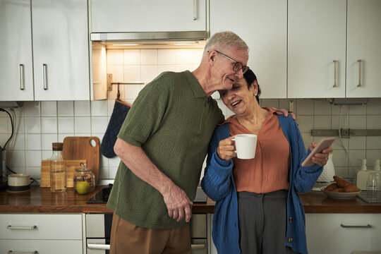 Elderly couple standing in kitchen, smiling while having morning coffee and using smartphone. Senior lady holding mug, senior man embracing affectionately