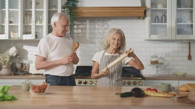 Enjoy simple life and be happy, old wife and husband dancing in home kitchen. Elderly lady singing into rolling-pin, aged man dancing with wooden spoon, fun and joy, merry weekend of senior spouses