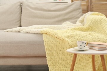 Comfortable sofa with woolen blanket, books and cup on side table indoors
