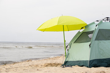 A green tent with a yellow umbrella stands on a sandy beach by the sea. A peaceful seaside camping...