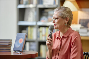 Caucasian senior woman speaking into microphone during book event, sitting at table with stack of books and book cover displayed, wearing glasses and earrings, bookshelves in background