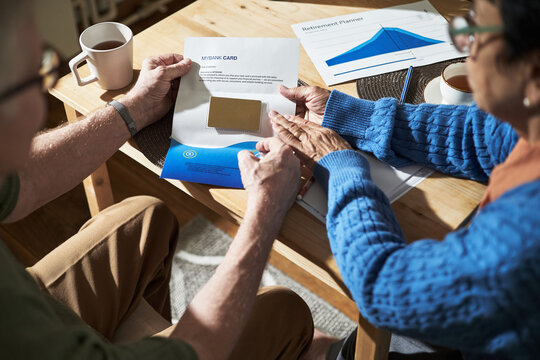 Senior couple opening envelope with credit card, while reviewing retirement plans at home, with table and documents in background, creating a moment of shared decision-making