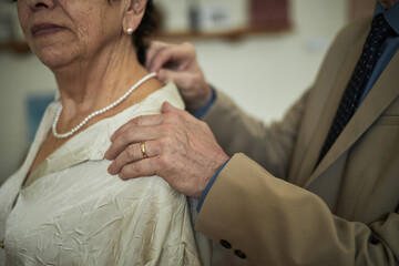 Hands of man adjusting necklace on elderly woman while she is standing in indoor setting with...