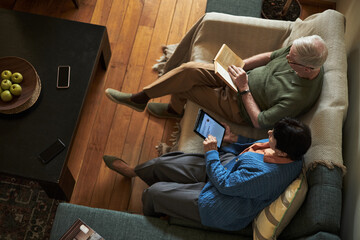Senior couple spending quality time sitting together on a sofa, man reading book while woman using...