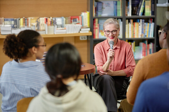 Senior Caucasian woman holding microphone speaking to diverse group of young adults and teenagers seated in library setting during session, bookshelves filled with books visible in background