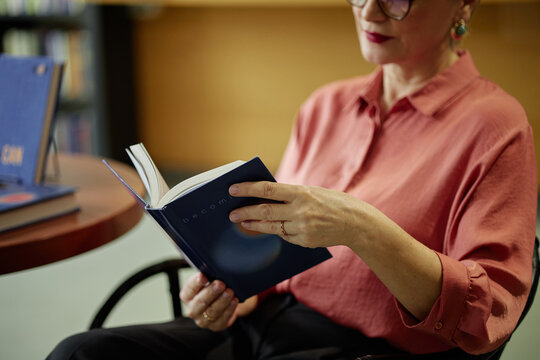 Middle aged Caucasian woman sitting and reading book with focused expression, holding open pages in hands, wearing glasses, bookshelf and table visible in background