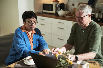 Elderly couple examining utility bills together while sitting at kitchen table with laptop and coffee cups, surrounded by household items