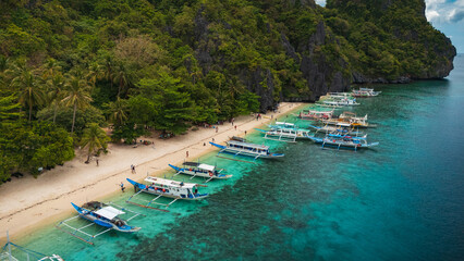 Aereal view of Colorful Bangka Boats Docked on the Crystal-Clear Waters and Coral Reefs of Entalula Island with a beautiful beach with people in El Nido, Palawan, Philippines
